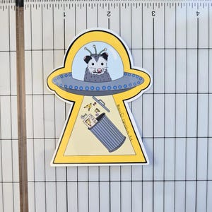 May include: A yellow and blue sticker featuring a possum in a UFO, with the text "Race Car Opossum Art" and a bin with pizza and other food scraps.