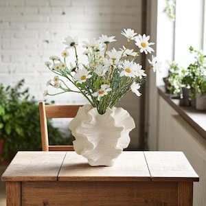 May include: A white ceramic vase with a ruffled, organic shape, filled with a bouquet of white daisies.