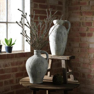 Könnte beinhalten: Zwei weiße Keramikvasen mit rustikalem Finish, eine hoch und eine niedriger, mit einem getrockneten Blumenarrangement in der niedrigeren Vase. Die Vasen stehen auf einem Holztisch und einem Hocker.