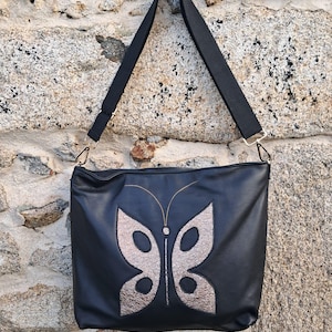 May include: A black leather shoulder bag with a silver butterfly design. The bag has a long adjustable strap.