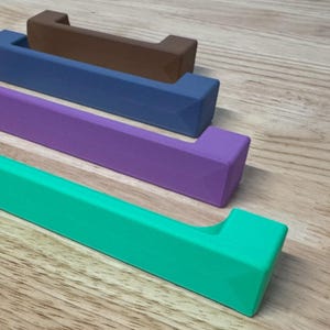 May include: A selection of colourful cabinet pulls in teal, purple, blue, and brown. The pulls are rectangular with a curved handle, and are arranged on a wooden surface. These decorative hardware pieces are for home decor.