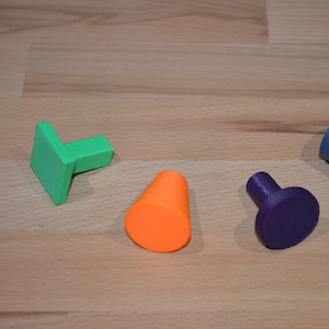 Op de afbeelding: Vier kleurrijke, geometrische vormen: een groene L-vorm, een oranje kegel, een paarse knop en een blauwe cilinder. De objecten zijn gerangschikt op een lichtbruin houten oppervlak. De vormen zijn solide en lijken 3D-geprint.