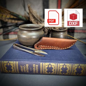 Puede incluir: Una funda de cuchillo de cuero marrón con un borde cosido, un pequeño par de tijeras de plata y una olla de cerámica marrón sobre un libro azul titulado "Grandes esperanzas" de Charles Dickens.