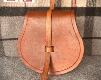 May include: A brown leather bag with a strap and a flap closure. The bag is on a plaid fabric background.