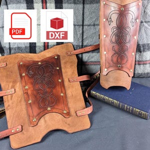 Puede incluir: Dos protectores de brazo de cuero marrón con nudos celtas decorativos y remaches de latón. Los protectores están sobre una superficie azul oscuro con un libro.