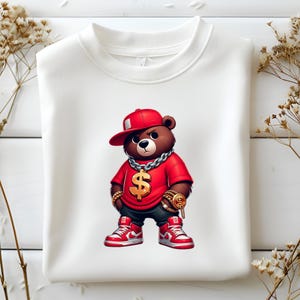 Puede incluir: Una sudadera blanca con una imagen de dibujos animados de un oso marr&oacute;n que lleva una gorra de b&eacute;isbol roja, una camiseta roja, una cadena de oro y un colgante de oro con forma de d&oacute;lar. El oso lleva zapatillas rojas.