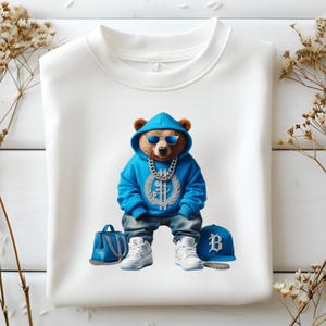 Puede incluir: Sudadera blanca con un oso azul que lleva gafas de sol, una cadena de oro, una sudadera con capucha azul, pantalones vaqueros azules, zapatillas blancas, un bolso de mano azul y una gorra de b&eacute;isbol azul con una B blanca.