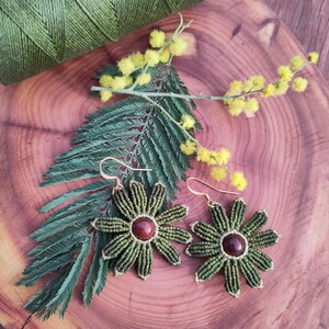 Peut inclure: Boucles d'oreilles fleurs en macramé vert foncé et or. Chaque boucle d'oreille présente une pierre centrale rouge foncé et des crochets dorés. Elles sont présentées sur une surface en bois avec du fil vert, des fleurs jaunes et des feuilles vertes.