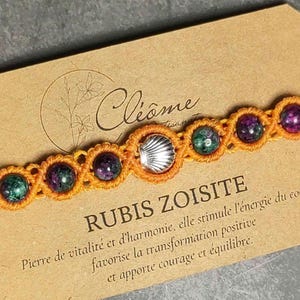 Peut inclure: Un bracelet macramé orange orné de perles de rubis zoisite et d'un charm en forme de coquillage argenté. Le bracelet est présenté sur une carte avec du texte en français, dont "RUBIS ZOISITE".