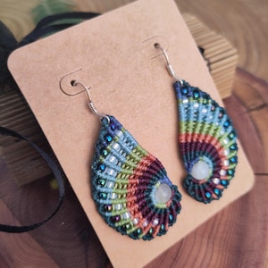 Peut inclure: Une paire de boucles d'oreilles faites à la main en forme de larme. Les boucles d'oreilles sont faites avec des fils et des perles colorés, et présentent une pierre blanche au centre. Les boucles d'oreilles sont suspendues à des crochets argentés.
