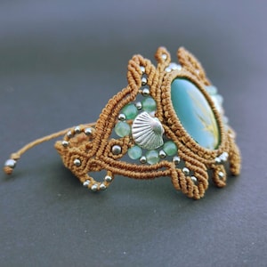Op de afbeelding: Een bruine macramé armband met een groene edelsteen en zilveren accenten. De armband is versierd met kleine groene kralen en een zilveren schelpbedel.
