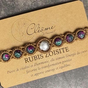 Può includere: Braccialetto in macramè con perline di rubino zoisite verdi e viola, con un ciondolo a forma di conchiglia argentata. Il braccialetto è esposto su una carta marrone con testo in francese.