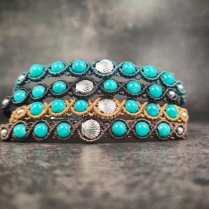Puede incluir: Cuatro pulseras de macramé con cuentas turquesas y detalles de conchas plateadas. Las pulseras son en tonos turquesa, negro, beige y marrón. Las pulseras están apiladas una encima de otra.
