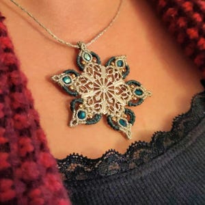 Peut inclure: Un collier pendentif flocon de neige argenté avec des accents turquoise et dorés. Le design en filigrane complexe présente des pierres précieuses de couleur turquoise. Le collier est porté sur un haut bleu foncé avec une bordure en dentelle et une écharpe texturée rouge et violette.