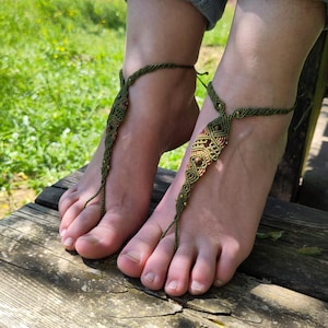 Può includere: Sandali a piedi nudi in macramè verde fatti a mano. I sandali presentano un'intricata annodatura con piccole perline color oro e un anello per le dita. I sandali sono indossati su una superficie di legno, con uno sfondo di erba verde e jeans.