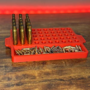 May include: A red plastic ammunition tray filled with brass casings and bullets. The tray has multiple holes to hold the casings upright, with a section for loose bullets. The background has a red glow.