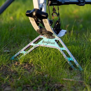 May include: A teal and red BowStruck bow stand is set in a grassy field. The stand has a three-legged design with the brand name printed on the front. The bow is white and black.