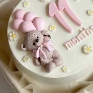 Op de afbeelding: Een ronde, witte taart versierd met een lichtbruine teddybeer die roze ballonnen vasthoudt. De taart is versierd met het roze nummer "11" en het woord "months" in roze. Kleine witte en gele bloemdecoraties zijn over de taart verspreid.