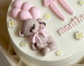 May include: A round, white cake decorated with a light brown teddy bear holding pink balloons. The cake is adorned with pink number "11" and the word "months" in pink. Small white and yellow flower decorations are scattered around the cake.