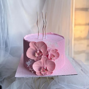 May include: A pink frosted cake decorated with three pink orchid flowers and three wavy gold candles. The cake sits on a square pink base and is displayed on a gold stand. The background is a sheer white fabric.