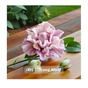 Peut inclure: Gros plan d'un moule en silicone de fleur d'iris violet clair avec le texte "IRIS Silicone Mold". La fleur a plusieurs couches de pétales et est entourée de feuilles et de bourgeons verts. Le moule est sur une surface en bois.