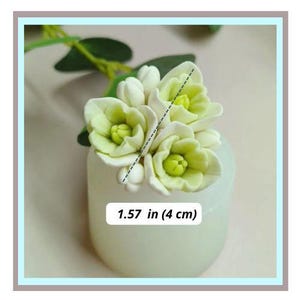 Puede incluir: Un pequeño jarrón blanquecino con un grupo de flores blancas y verde claro. Las flores tienen un diseño de pétalos detallado y en capas. La imagen incluye el texto "1.57 in (4 cm)", que indica la altura del arreglo.