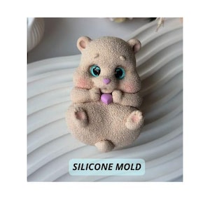 May include: A beige silicone mold in the shape of a cartoon bear holding a purple heart. The bear has large blue eyes, pink cheeks, and small ears. The text "SILICONE MOLD" is printed on a light blue rectangle.