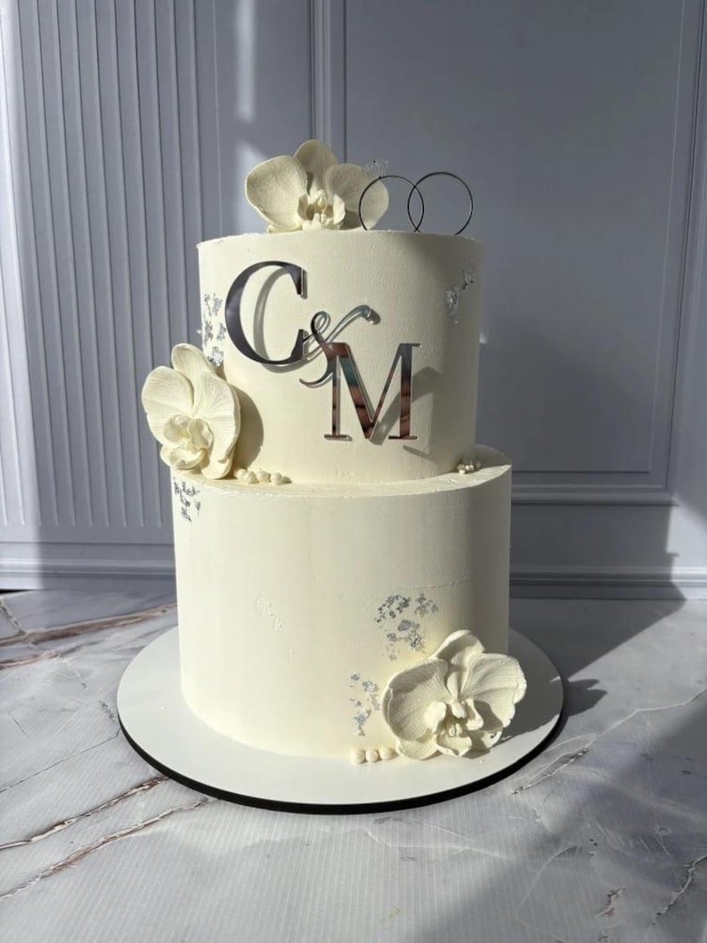 May include: Two-tiered white wedding cake decorated with white orchids and silver accents. The cake features the initials "C&M" in silver and a silver ring cake topper. The cake sits on a white cake board.