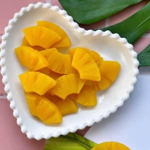 May include: A white heart-shaped dish with a scalloped edge filled with bright yellow pineapple slices. The fruit pieces are cut into wedges and arranged in a pile. A single pineapple ring is visible at the bottom edge of the frame.