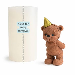 May include: A light-colored cylindrical candle with a dotted line and the text "A cut for easy removal." Next to it is a brown teddy bear figurine wearing a gold party hat.