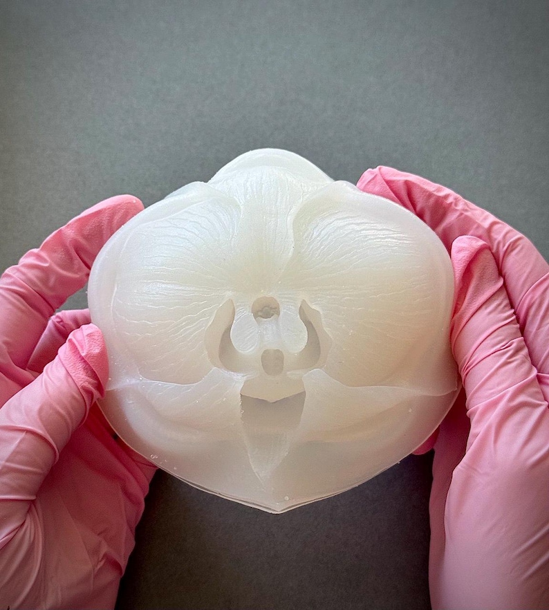 May include: A translucent, off-white orchid-shaped mold held by hands wearing pink gloves. The mold has detailed petal and stem impressions, and a flat base. The background is a solid gray.