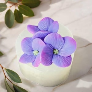 May include: A white, oval soap bar decorated with three purple and pink flower blossoms. The flowers have green centres and are arranged on top of the soap. The background is a marble surface with green leaves.