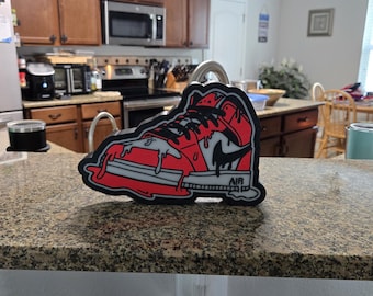 May include: A red, black, and gray sneaker-shaped patch with a dripping paint design. The patch features a black Nike swoosh and the word "AIR" on the sole. The patch is placed on a speckled countertop.