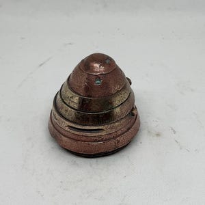 May include: A small, metallic decorative object with a layered design. The object has a copper-coloured top and alternating bands of copper and gold. The overall shape is rounded and conical, with a slightly textured surface.