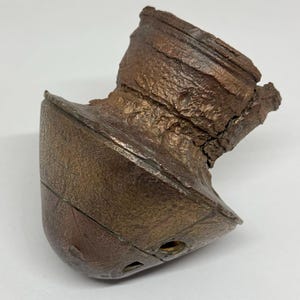 May include: A weathered, copper-coloured ceramic pot with a unique, asymmetrical shape. The pot has a wide, open top and a narrow, pointed base. The surface of the pot is rough and textured, with visible cracks and imperfections.