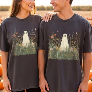 May include: Dark grey t-shirts featuring a whimsical ghost design in a field of wildflowers. The ghost is white with black eyes, and the flowers are in shades of orange, pink, and yellow. The shirts are a relaxed fit.