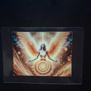 Könnte beinhalten: Ein rechteckiger Aufkleber mit einer zentralen Figur, die große, weit ausgebreitete goldene Flügel hat. Die Figur ist von strahlendem Licht umgeben und unter dem Oberkörper befindet sich ein markantes goldenes Spiralmuster auf dunklem Hintergrund.