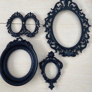 May include: Four black ornate picture frames in various shapes: oval, round, and double oval. The frames have detailed scrollwork and decorative elements. The frames are displayed on a light wood surface.