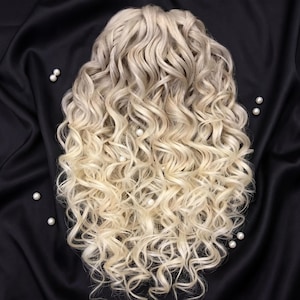 Op de afbeelding: Blonde, krullende pruik met een gradiënteffect, van een donkerdere aanzet naar een lichtere, bijna platina blonde kleur aan de uiteinden. De pruik is gestyled met volumineuze, veerkrachtige krullen en staat op een zwarte satijnen achtergrond, met verspreide parels.
