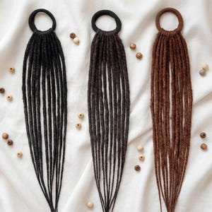 Puede incluir: Tres extensiones de rastas en negro, marrón oscuro y marrón claro, cada una unida a una goma para el pelo negra o marrón. Pequeñas cuentas de madera están esparcidas alrededor de las extensiones sobre un fondo de tela blanca. Las rastas son largas y rectas.