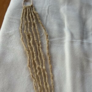 May include: A close-up of a beige macrame piece with multiple strands. The strands are knotted together at the top, forming a loop. The macrame is set against a white, textured background.