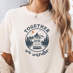 May include: Ivory-colored t-shirt with a circular graphic featuring a camper van on a road, mountains, and trees. The text reads "TOGETHER we wander est. 2020." The shirt is a relaxed fit.