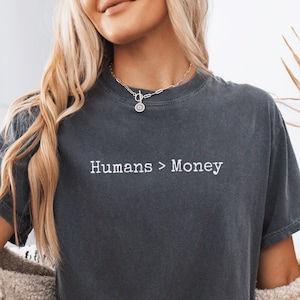 May include: Dark grey t-shirt with the text "Humans > Money" in white. The shirt has short sleeves and a crew neck. The word "Pepper" is at the bottom right corner of the image.