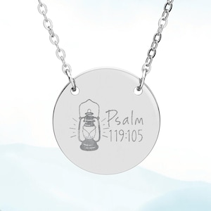 May include: A silver stainless steel necklace with a circular pendant. The pendant has an engraved lantern design and the text "Psalm 119:105". The necklace is set against a light blue and white backdrop. The words "Strength and Song" and "Stainless Steel" are also visible.