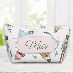 Puede incluir: Bolsa de cosméticos blanca con cremallera, con un estampado de acuarela de bailarinas, cascanueces y elementos botánicos. Una etiqueta rosa muestra el nombre "Mia". La bolsa está sobre una superficie de madera blanca.