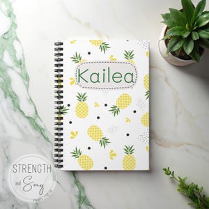 May include: A spiral-bound notebook with a white cover featuring a repeating pattern of yellow pineapples, black dots, and yellow floral accents. The name "Kailea" is displayed in a green oval. A small plant is in the background.