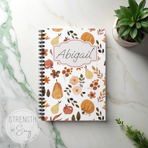 Puede incluir: Un cuaderno con espiral con un diseño de temática otoñal que presenta calabazas, peras y elementos florales. El nombre "Abigail" se muestra en un marco decorativo. El cuaderno está sobre una superficie de mármol, con una pequeña planta en maceta en el fondo.