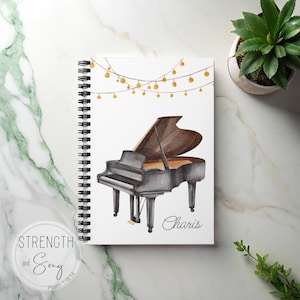 May include: Spiral-bound notebook with a watercolor illustration of a grand piano. The piano is black and brown, with the name "Charis" written below it. A string of golden lights hangs above the piano. The notebook is on a marble surface.