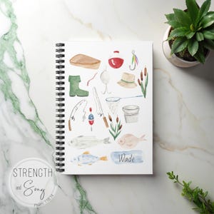 May include: A spiral-bound notebook with a watercolor illustration of fishing-related items. The design includes a fishing rod, fish, a bucket, a hat, and the name "Wade". The notebook is on a marble surface with a plant in the background. The text "Strength and Song" is on the bottom left.