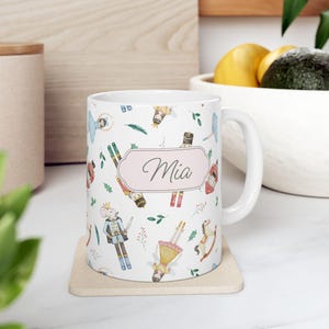 May include: White ceramic mug with a repeating pattern of watercolor illustrations of nutcrackers, fairies, and other holiday-themed elements. The name "Mia" is displayed in a pink oval. The mug sits on a beige coaster.